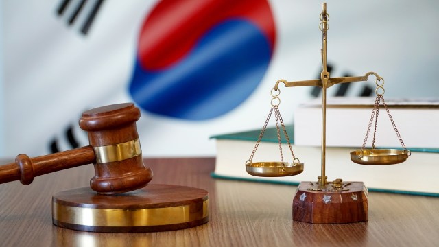 A gavel and balance scales on a table with a blurred South Korean flag in the background.