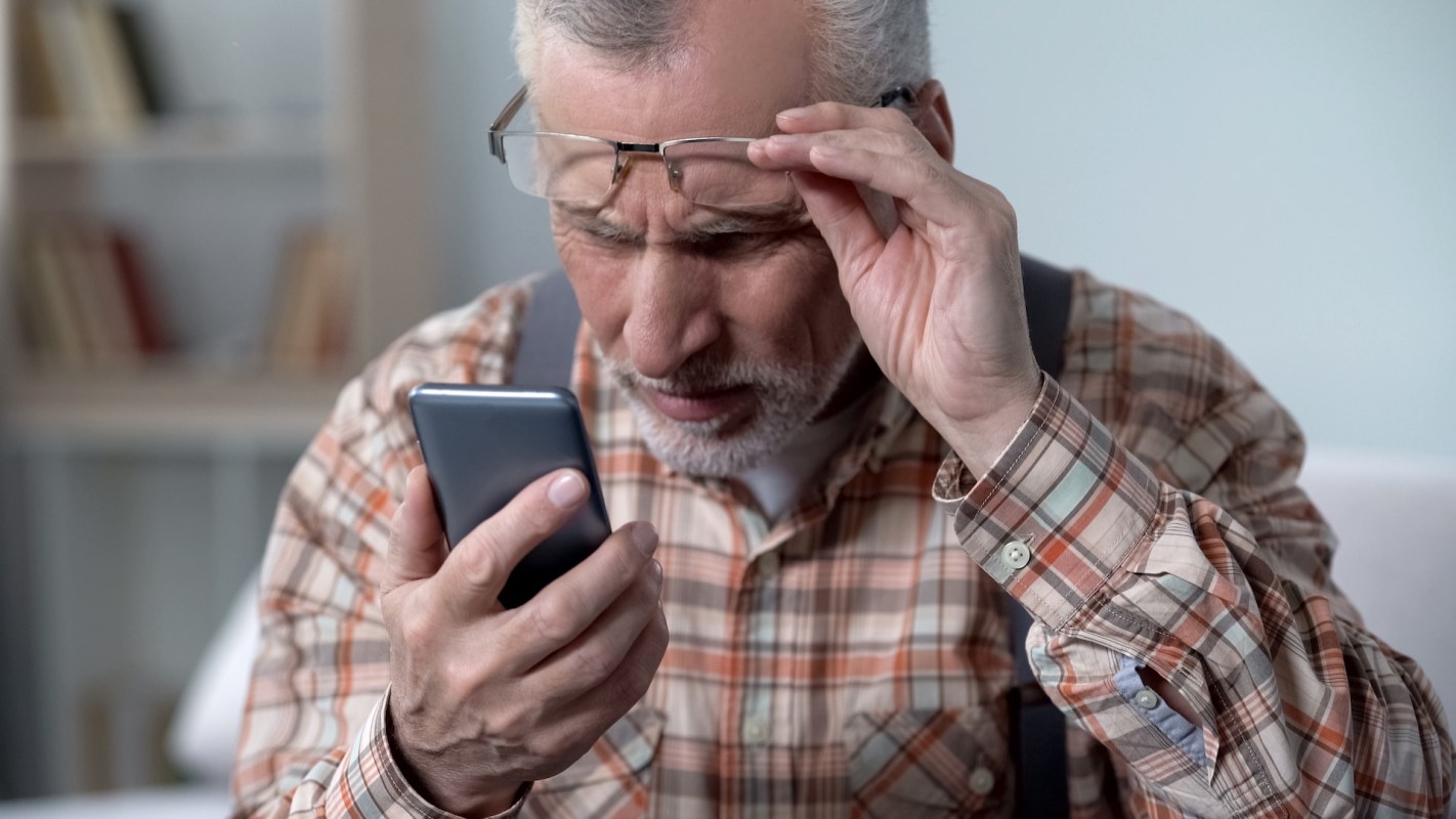 An elderly man adjusts his glasses, looking confused, while looking at a smartphone.