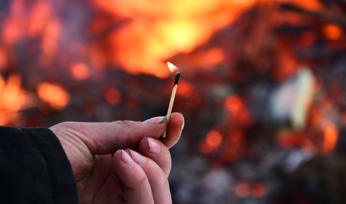 A hand holds a matchstick with a small flame against a blurred background of burning embers.