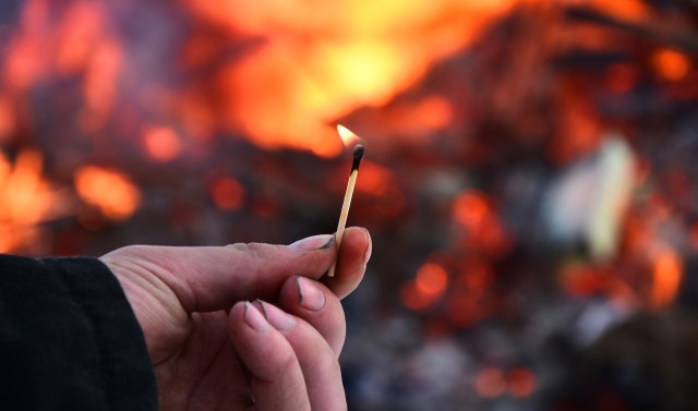 A hand holds a matchstick with a small flame against a blurred background of burning embers.