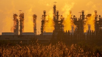 A factory emitting steam against an orange sunset, with tall grass in the foreground.