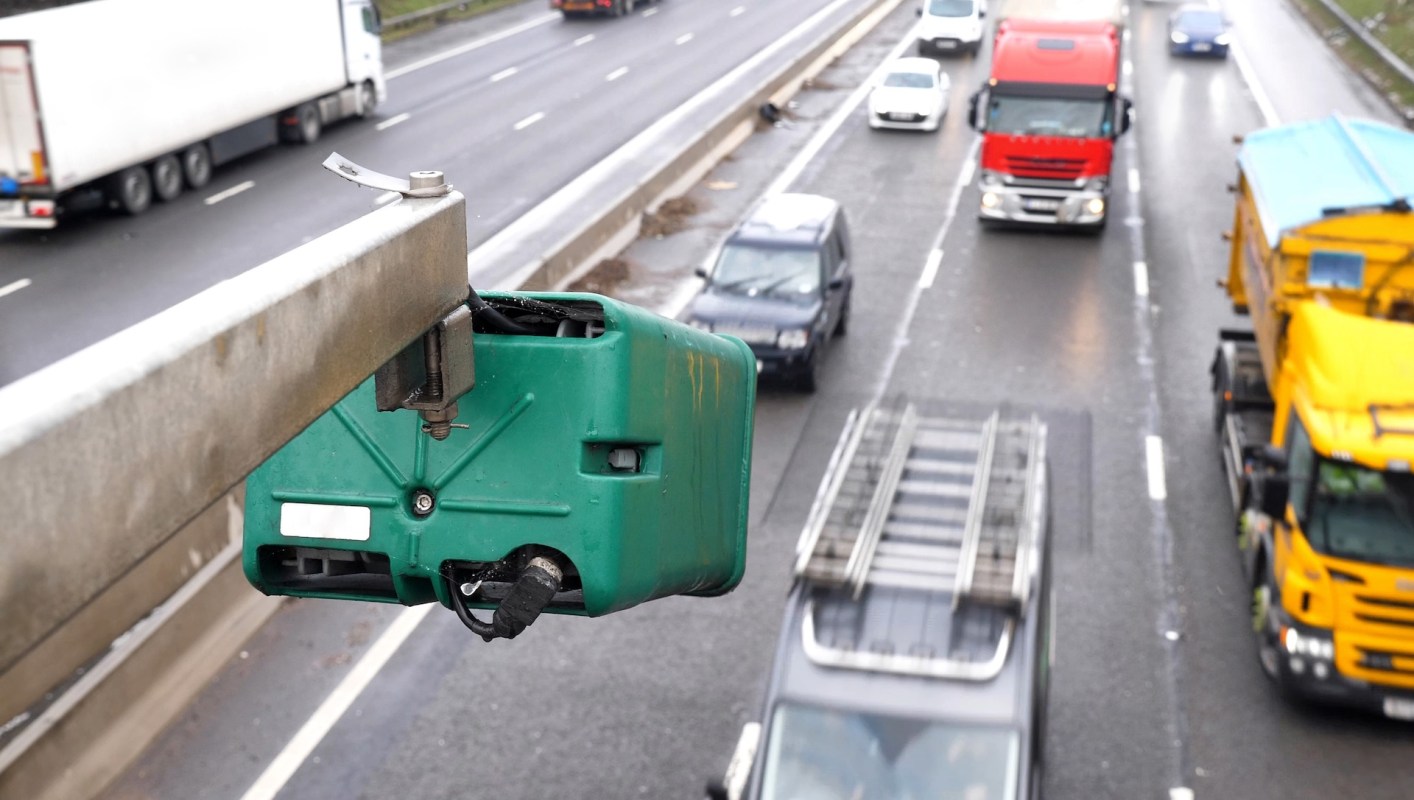 A green camera mounted on a pole above a busy highway with various vehicles in motion below.