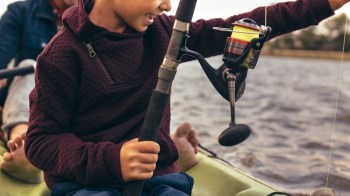 A child holding a fishing rod on a kayak with water in the background.