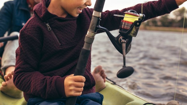 A child holding a fishing rod on a kayak with water in the background.