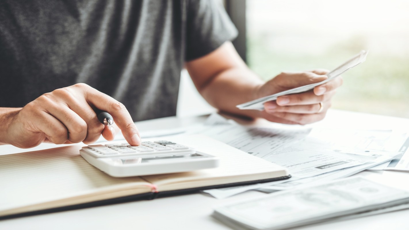 A person calculates expenses using a calculator and holds bills in their other hand, surrounded by documents.