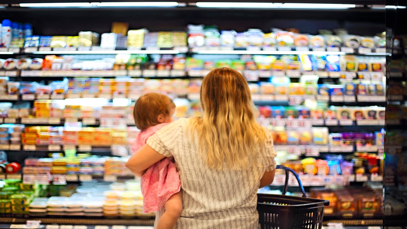 A woman holds a child while browsing refrigerated food items in a grocery store.