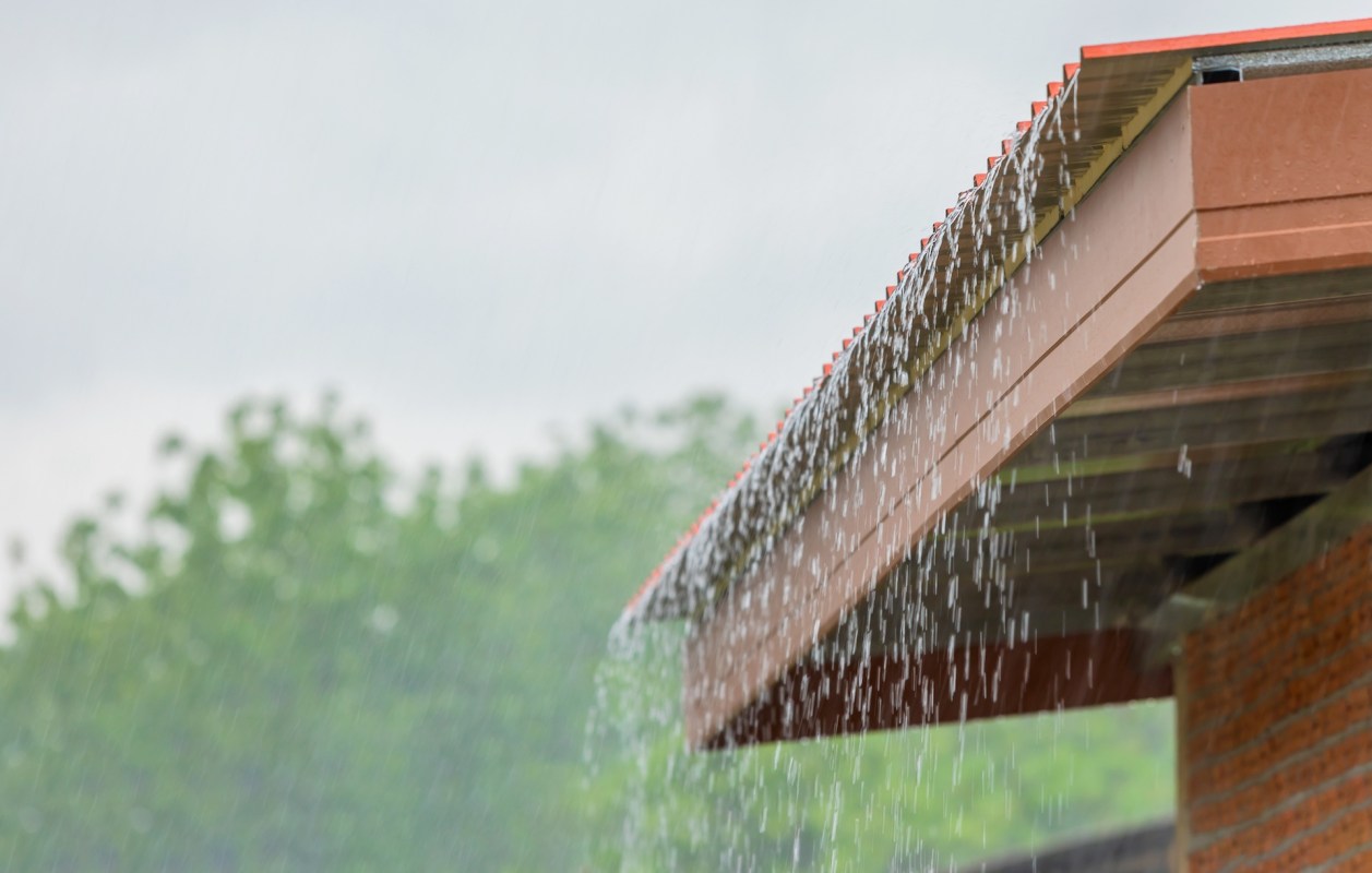 Water cascades from the edge of a roof during rain, with blurred greenery in the background.