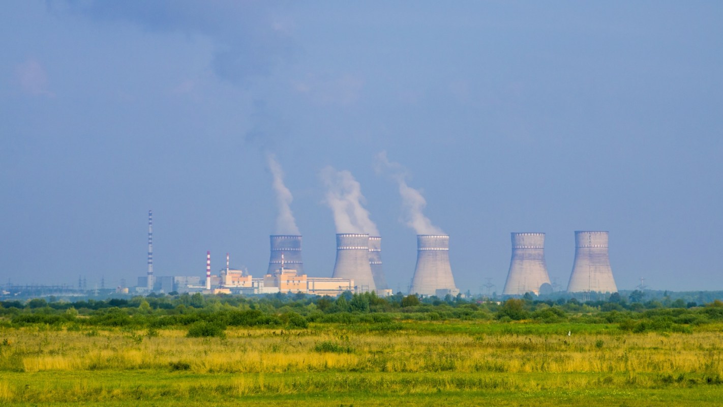 A nuclear power plant with cooling towers emitting steam, set against a green landscape.