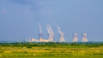 A nuclear power plant with cooling towers emitting steam, set against a green landscape.