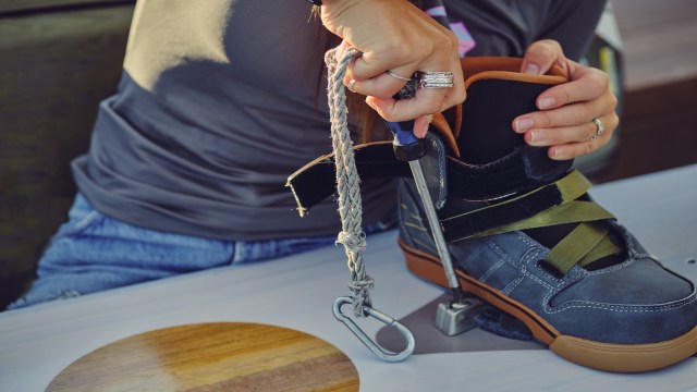 A hand holds a rope while adjusting a shoe with a clip on a table with a circular wooden surface.