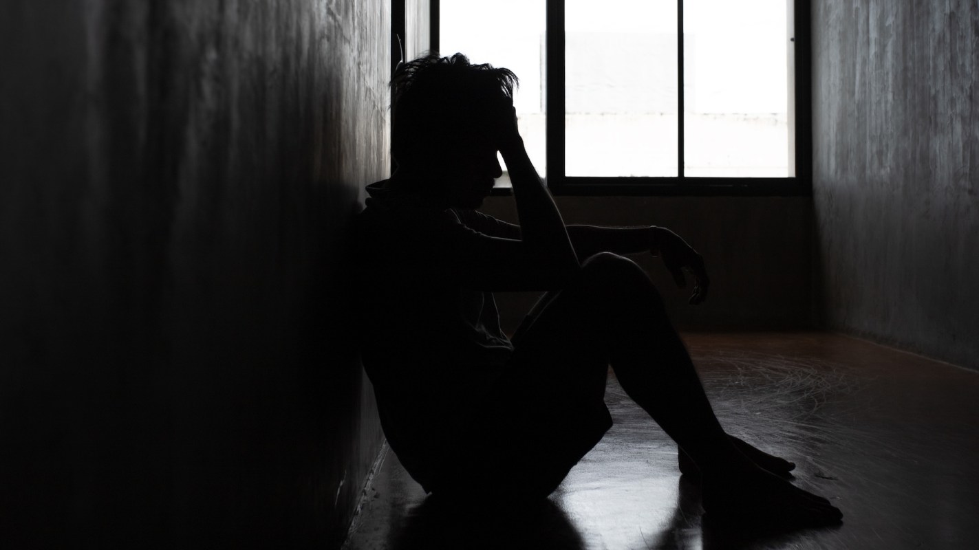 A person sits against a wall in a dimly lit room with a window in the background.
