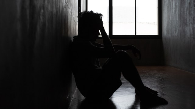A person sits against a wall in a dimly lit room with a window in the background.