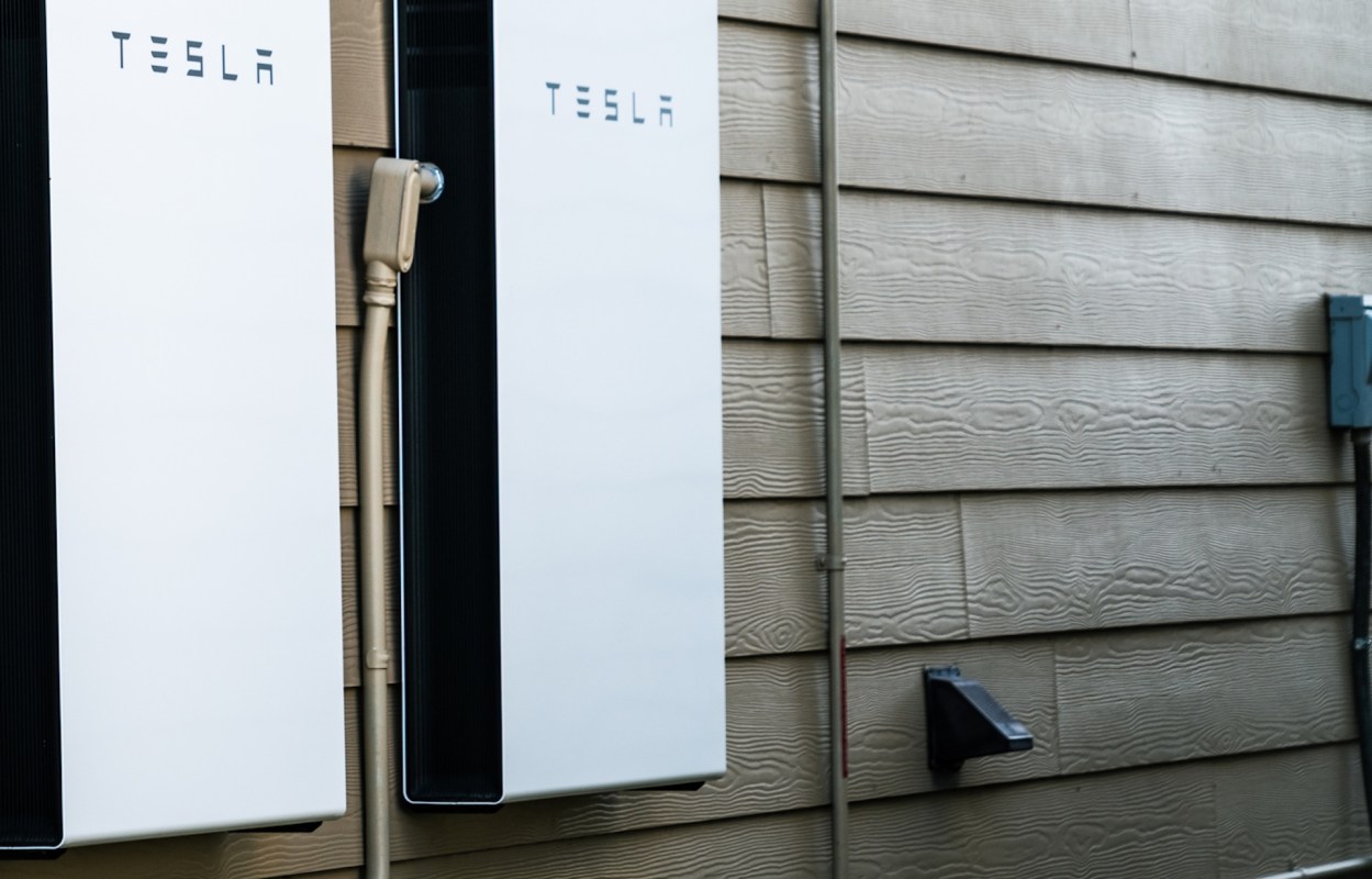 Two Tesla Powerwall energy storage units mounted on a textured wall with electrical connections and a nearby electrical box.