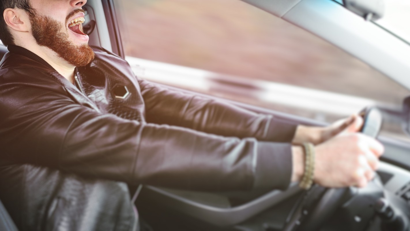 A man wearing a leather jacket is driving a car while yelling.