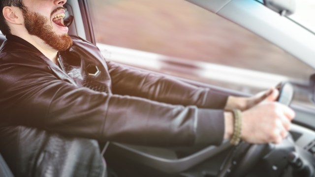 A man wearing a leather jacket is driving a car while yelling.