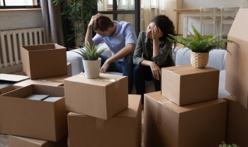 A man and a woman sit with their hands on their heads amid unpacked moving boxes in a living room.