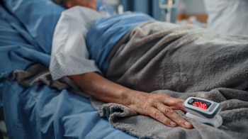 A hand rests on a blanket in a hospital bed, with a pulse oximeter displaying readings.
