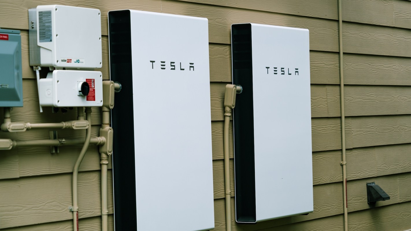 Two Tesla Powerwall units mounted on a wooden wall, with electrical components and pipes visible nearby.