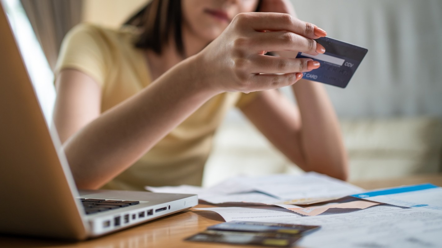 A woman holds a credit card while sitting at a table with a laptop and various papers.