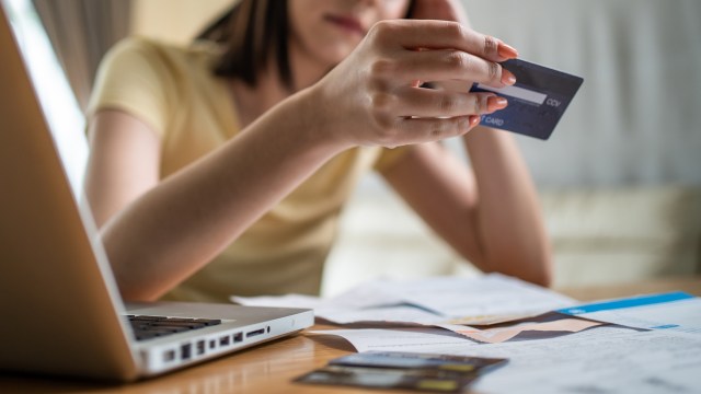 A woman holds a credit card while sitting at a table with a laptop and various papers.