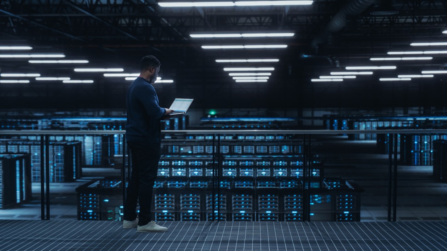A person stands in a data center using a laptop, surrounded by rows of servers.