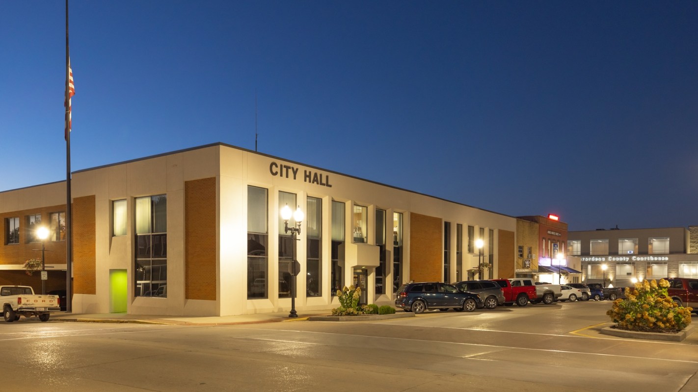 A city hall building lit up at night, with parked cars and streetlamps in the foreground.
