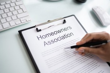 A hand holding a pen is positioned over a clipboard with a document titled "Homeowners Association."
