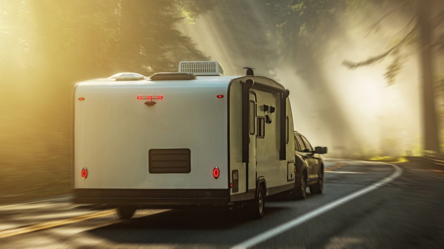 A vehicle towing a camper travels along a winding road in a forested area.