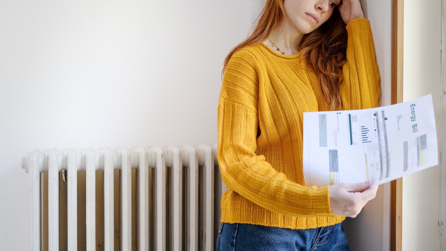 A woman holds a printed energy bill in disbelief while leaning against a wall next to a radiator.