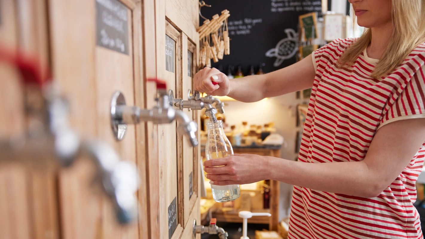 A person fills a glass bottle from a tap at a wooden beverage dispenser.