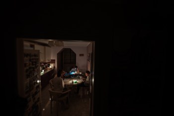 A family gathers around a table with glowing candles and a laptop in a dimly lit kitchen.
