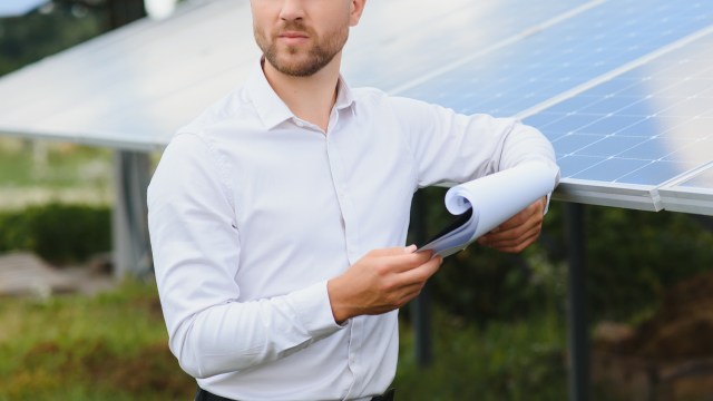 A man holds a rolled document while standing next to solar panels in a grassy area.