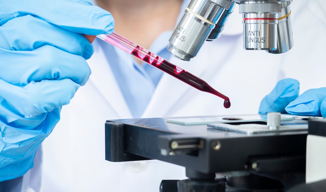 A person in a lab coat uses a pipette to transfer blood onto a microscope slide.