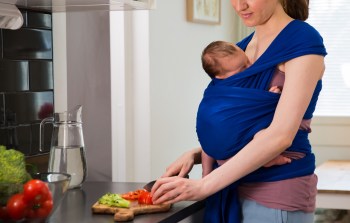 A woman cutting vegetables while holding a baby in a sling.