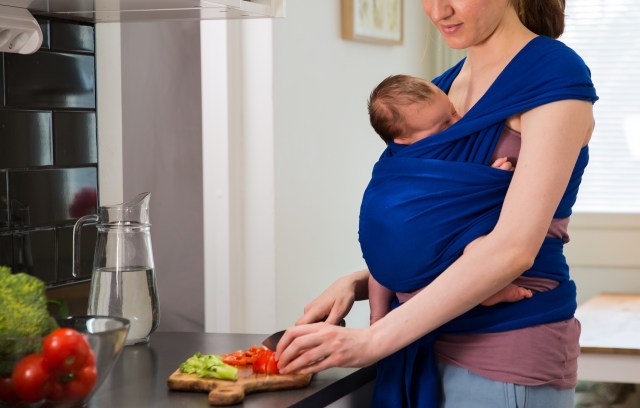 A woman cutting vegetables while holding a baby in a sling.