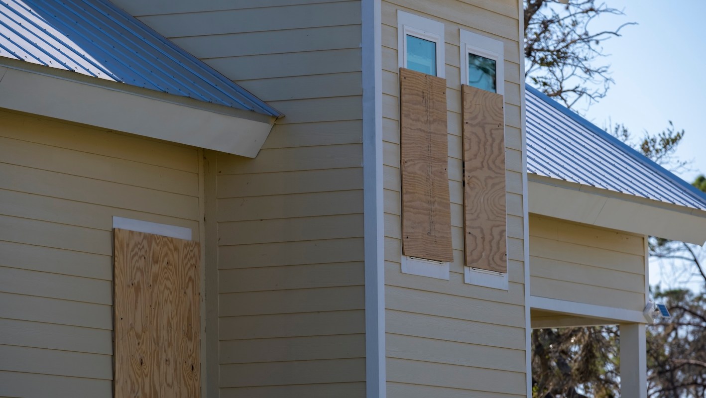 A house with plywood boards covering two windows on a light-colored exterior.