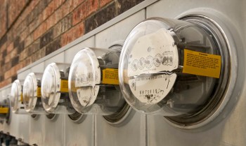Five electric meters mounted on a wall with a brick background.