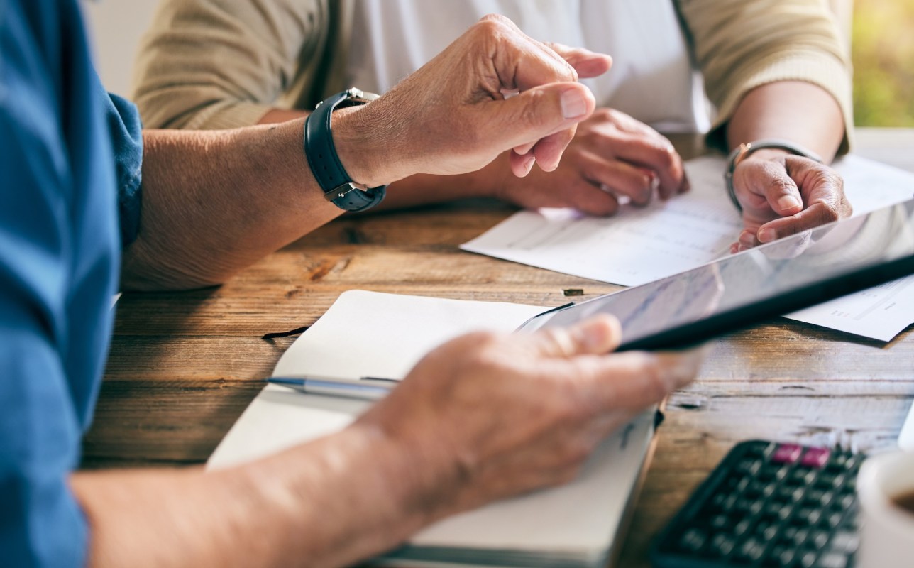 Two people discuss documents on a table with a calculator, one pointing at a tablet screen and the other holding papers.