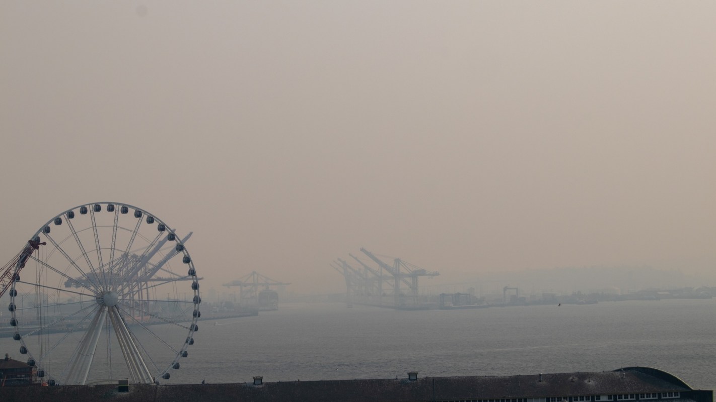 A ferris wheel stands near a hazy Seattle harbor with cranes and boats in the distance.
