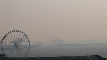 A ferris wheel stands near a hazy Seattle harbor with cranes and boats in the distance.