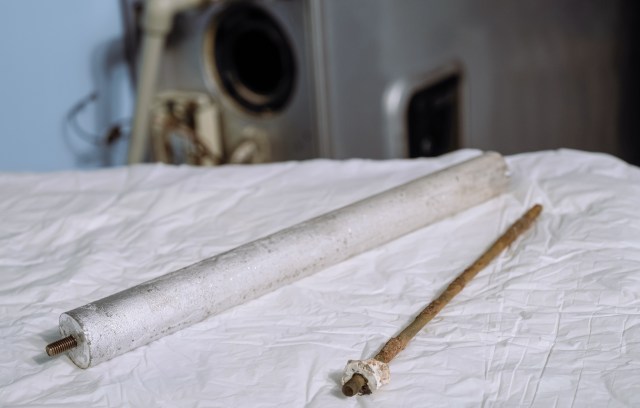 Two cylindrical rods, one silver and one brown, resting on a white surface with a blurred water heater in the background.
