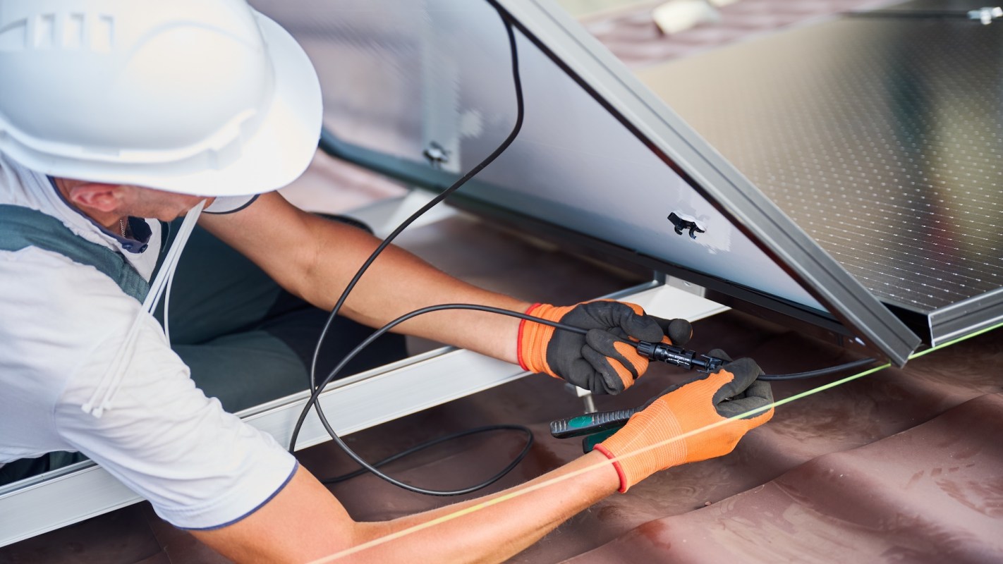 A worker connects wires while installing a solar panel on a rooftop.