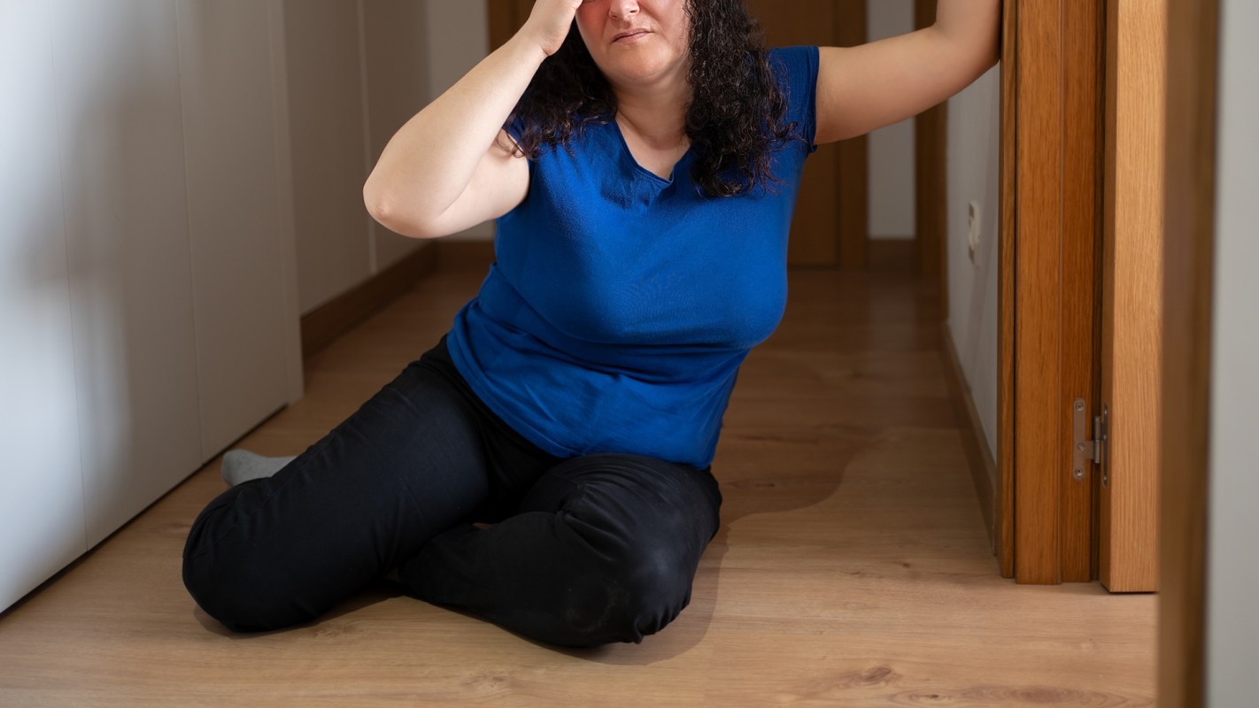 A woman kneels in a hallway, resting her head against the wall with one hand on her forehead.