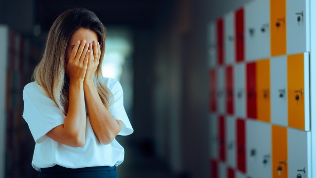 A teacher covers her face with her hands in a corridor with lockers.
