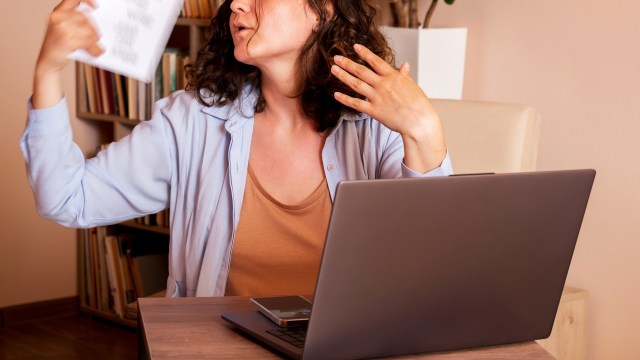 A woman gestures while sweating at a desk with a laptop and a mobile phone.