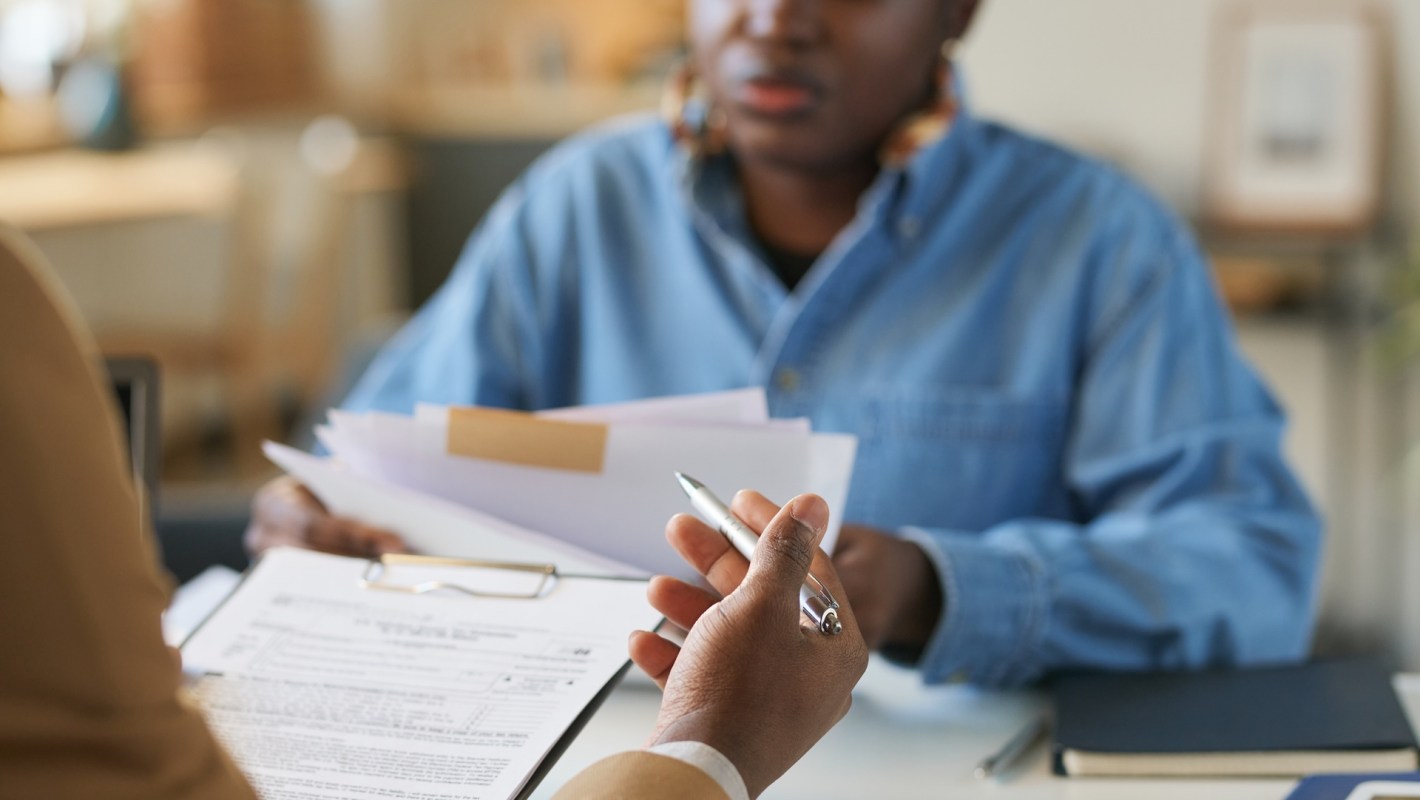 A person holds a pen and paperwork while another person hands over documents in an office setting.