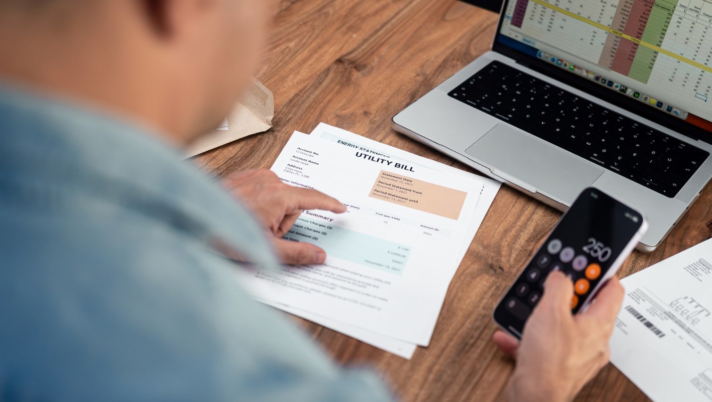 A person reviews a utility bill while using a calculator on a smartphone, with a laptop and documents on the table.