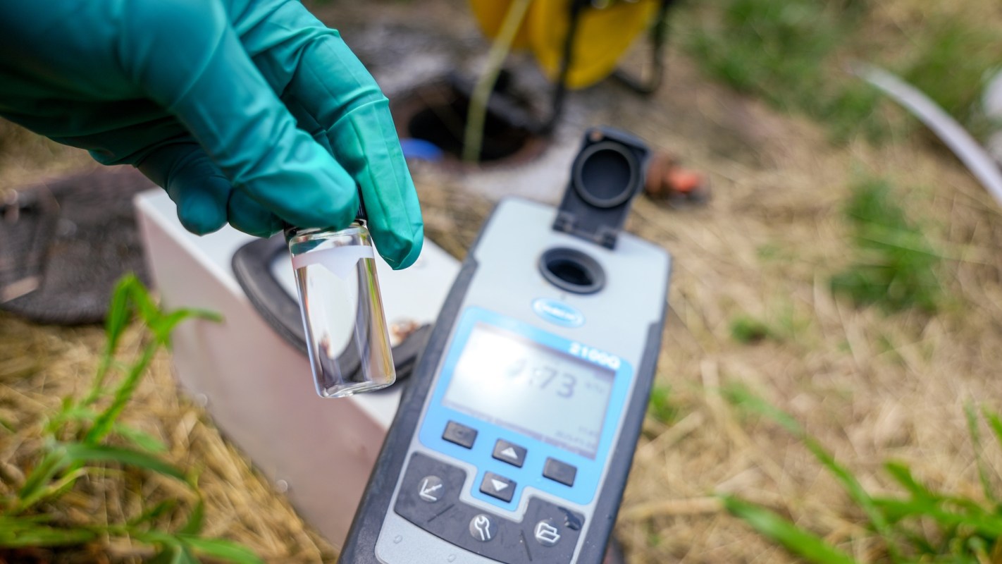 A person in gloves holds a test tube with a liquid near a portable measurement device displaying a reading.