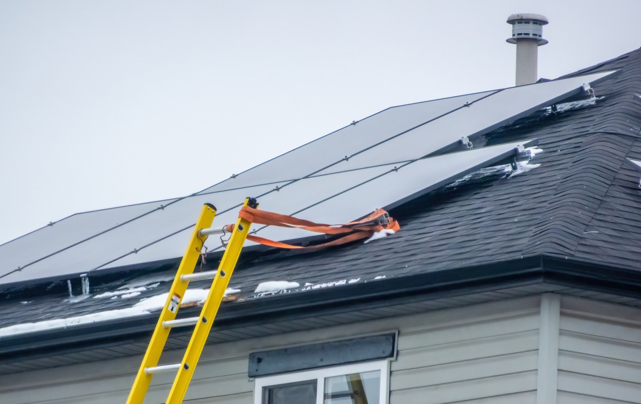 A yellow ladder is leaning against a house with solar panels partially covered in snow.