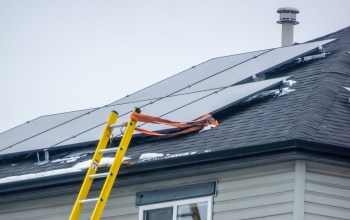 A yellow ladder is leaning against a house with solar panels partially covered in snow.